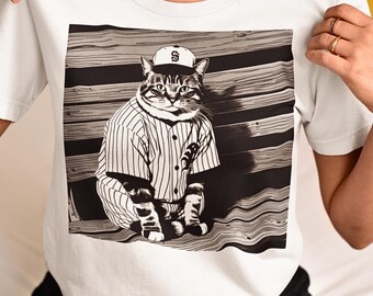 May include: White t-shirt with a sepia-toned image of a cat in a baseball uniform. The cat wears a cap with an "S" and a pinstriped jersey. The cat is sitting on a wooden bench, creating a unique and playful design.