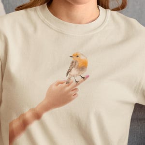 May include: Cream-colored t-shirt featuring a watercolor illustration of a robin perched on a finger with pink nail polish. The bird has orange and brown feathers. The design is centered on the shirt.
