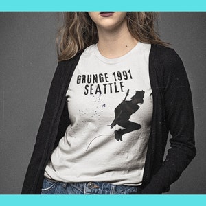 Puede incluir: Una camiseta blanca con una silueta negra de una persona tocando una guitarra. El texto "GRUNGE 1991 SEATTLE" está impreso en la camiseta.