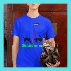 Puede incluir: Una camiseta azul con un gráfico de dos gatos negros persiguiendo un rayo láser rojo de un platillo volante. El texto "Creatures are fun" está impreso debajo del gráfico. Una persona está sosteniendo un gato atigrado en sus brazos.