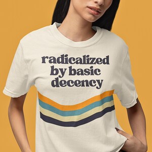 May include: A white t-shirt with the text "radicalized by basic decency" in black lettering. The shirt features a retro-style striped design in blue, yellow, and white.