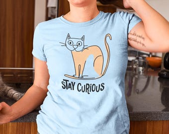 Pode incluir: Uma camiseta azul claro com uma ilustração de desenho animado de um gato surfando. O texto "STAY CURIOUS" é impresso abaixo do gato.