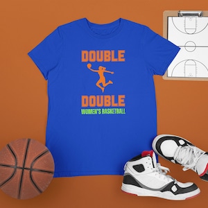 Puede incluir: Una camiseta azul real con el texto "DOUBLE DOUBLE BALONCESTO FEMENINO" y una silueta de una jugadora de baloncesto en naranja.