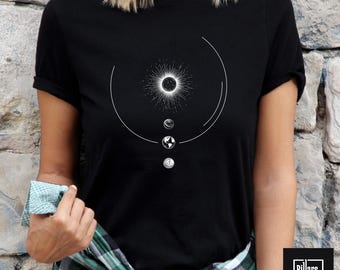 Może przedstawiać: Czarny t-shirt z białym nadrukiem przedstawiającym zaćmienie słońca i planety. Koszulka ma okrągły dekolt i krótkie rękawy. Tekst "Black" znajduje się w lewym górnym rogu.