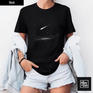 Puede incluir: Camiseta negra con un gráfico blanco de una estrella fugaz sobre un paisaje con un lago y montañas. La camiseta tiene cuello redondo y mangas cortas. El texto "Black" está en un rectángulo negro en la parte superior izquierda. El logotipo de la marca está en la parte inferior derecha.