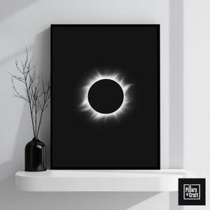 May include: A framed black and white print depicting a solar eclipse. The artwork features a dark central circle surrounded by a bright, radiant corona. A black vase with branches sits to the left on a white shelf. The print is labeled "Pillars of Craft".