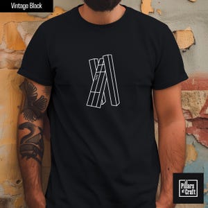 Pode incluir: Camiseta preta com um design geométrico branco. O design apresenta uma letra "A" estilizada composta por formas retangulares. O texto "TARS" é visível dentro do design. A camiseta é rotulada como "Vintage Black" e tem um logotipo "Pillars of Craft".