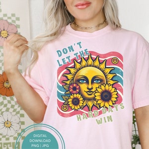 Op de afbeelding: Zachtroze T-shirt met een retro-afbeelding. Het ontwerp toont een lachende zon met zonnebloemen, blauwe ogen en de tekst "DON'T LET THE HARD DAYS WIN". De afbeelding bevat een vredesteken.
