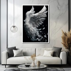 May include: A framed art print featuring a detailed depiction of a white angel wing against a black background. The wing is rendered with soft, feathery textures, with some feathers drifting away. The artwork is displayed in a modern living room setting.