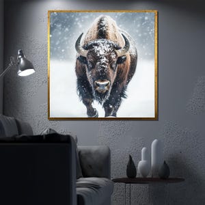 May include: A framed print of a bison in a snowstorm. The bison is brown and black with large horns, and is facing the viewer. The artwork is in a gold frame and is hanging on a gray wall.