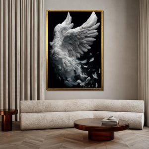 May include: A framed art print featuring a pair of white angel wings against a black background. The wings are detailed with individual feathers, some of which appear to be falling away. The frame is gold, and the artwork is displayed in a modern living room setting.