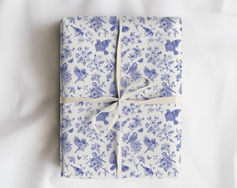 May include: A rectangular gift wrapped in white paper with a blue floral and bird pattern. The package is tied with a cream-colored ribbon. The design features birds, flowers, and butterflies.