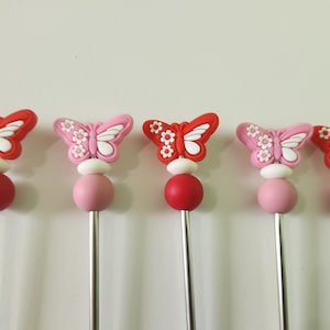 Może przedstawiać: Pięć dekoracyjnych szpikulców z motylami. Każdy szpikulec ma motyla w kolorze czerwonym lub różowym z białymi akcentami, białą kulkę, czerwoną lub różową kulkę i srebrny metalowy trzon. Idealne do przystawek lub koktajli.