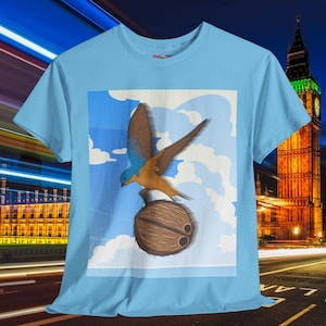 Könnte beinhalten: Hellblaues T-Shirt mit einer Cartoon-Illustration eines Vogels mit braunen und blauen Flügeln, der auf einer Kokosnuss sitzt. Der Hintergrund zeigt einen blauen Himmel mit weißen Wolken. Das Shirt hat einen Rundhalsausschnitt.