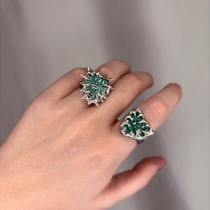 Puede incluir: Dos anillos plateados con detalles verdes y plateados. Uno tiene un diseño espinoso en forma de estrella, el otro es triangular. Ambos anillos presentan un centro verde con forma de placa de circuito.