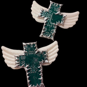 May include: Two decorative crosses with white glittery wings. The crosses are made from green circuit boards with silver edging. The background is black.