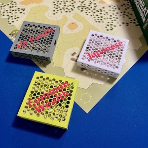May include: Three square game component boxes in gray, white, and yellow, each with a honeycomb pattern and the word "Japanese" in red. The boxes are on a game board with a blue surface.