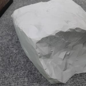 May include: A white, 3D printed rock with a textured surface. The rock has a rough, uneven shape and appears to be made of a lightweight material.