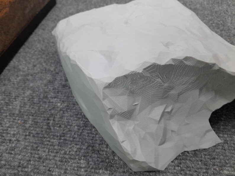 May include: A white, 3D printed rock with a textured surface. The rock has a rough, uneven shape and appears to be made of a lightweight material.