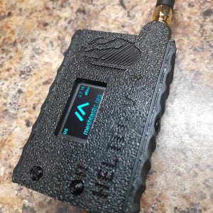 May include: Black handheld device with a textured surface, small screen displaying blue text, and a black antenna. The screen displays the text "meshtastic.org". The device has the word "HEL" printed on the side.