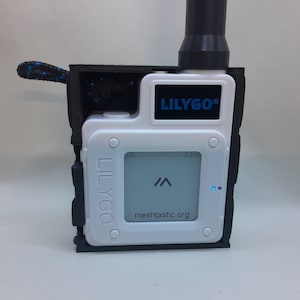 May include: A white LilyGo device with a black case and a black antenna. The device has a small screen that displays "meshcastic.org" and a blue LED light. The device is labeled "LilyGo" in blue text.