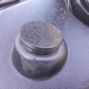 May include: Close-up of a metallic, textured, circular object with a flat top and a rounded base. The object is a dark gray color and appears to be made of metal. The surface it rests on is also dark gray and textured.