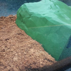 Pode incluir: Um close-up de um material verde amassado, semelhante a um tecido, que se assemelha a uma rocha, colocado sobre uma superf&iacute;cie marrom texturizada.