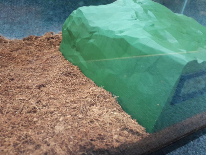 Pode incluir: Um close-up de um material verde amassado, semelhante a um tecido, que se assemelha a uma rocha, colocado sobre uma superf&iacute;cie marrom texturizada.