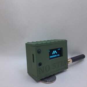 May include: A small, green, rectangular device with a black antenna and a screen displaying a blue arrow and the text "meshtactic.org". The device has the text "NO STEP" printed on the front.
