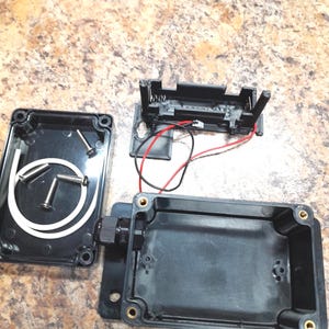 May include: An open black plastic project box with a black battery holder, screws, and a white rubber seal. Red and black wires are connected to the battery holder. The box has a black connector on one side.