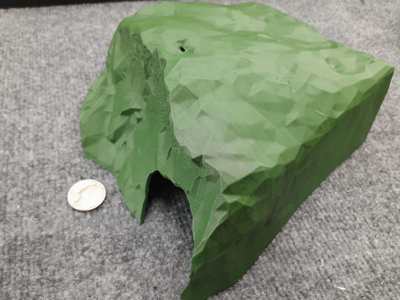 Pode incluir: Uma rocha verde, impressa em 3D, texturizada com uma abertura de caverna. A rocha &eacute; projetada para uso em layouts de trens em miniatura ou outras cenas em miniatura.