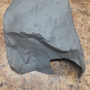 May include: A gray, 3D-printed rock formation with a textured surface and a cutout section. The rock has a rough, uneven appearance, with visible ridges and folds. The object is likely a decorative piece or a prop.