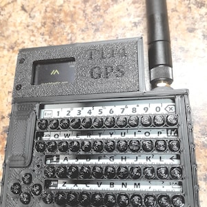 May include: A black 3D-printed GPS device with a keyboard layout and an antenna. The device has the text "T114 GPS" on the top and a small screen. The keyboard has a standard QWERTY layout with numbers and symbols.
