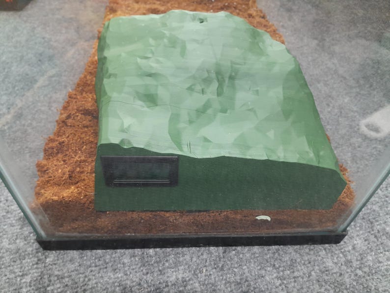 Pode incluir: Uma decora&ccedil;&atilde;o de terr&aacute;rio retangular em forma de rocha, de cor verde e texturada, com um term&ocirc;metro digital preto fixado no lado.