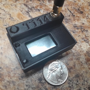 May include: Black electronic device with the text "T114" and a small screen. A quarter is next to the device for size comparison. The device has an antenna and appears to be made of plastic.