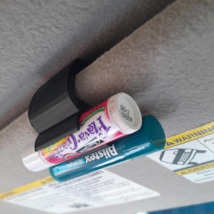 May include: A black plastic clip attached to a gray car visor, holding two cylindrical lip balm tubes. The top tube is white with pink and purple text, while the bottom tube is teal with white text. The clip secures both items firmly in place.