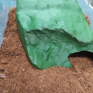 May include: A green plastic cave-like hide for small animals, such as reptiles or amphibians, placed on a bed of brown coconut coir substrate.