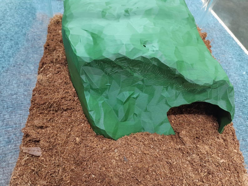 Pode incluir: Uma toca de pl&aacute;stico verde em forma de caverna para pequenos animais, como r&eacute;pteis ou anf&iacute;bios, colocada em uma cama de substrato de coco marrom.