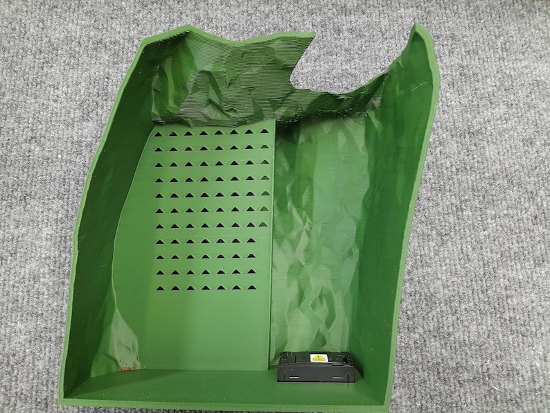 Pode incluir: Um recipiente de pl&aacute;stico verde com uma superf&iacute;cie texturizada e um pequeno objeto retangular preto dentro. O recipiente tem uma grade de pequenos tri&acirc;ngulos em um lado.