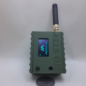 May include: A green, rectangular device with a small antenna, a screen displaying "mesh.tasic.org" and a small coin for scale.