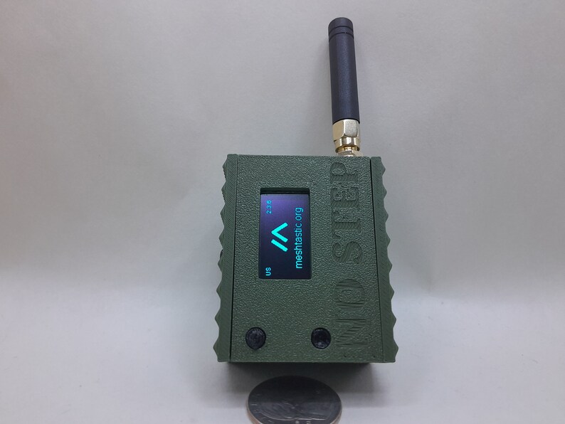 May include: A green, rectangular device with a small antenna, a screen displaying "mesh.tasic.org" and a small coin for scale.