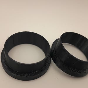 May include: Two black, circular objects with a raised, textured base. The objects are open cylinders, possibly components or parts. The items are set against a plain, off-white background.