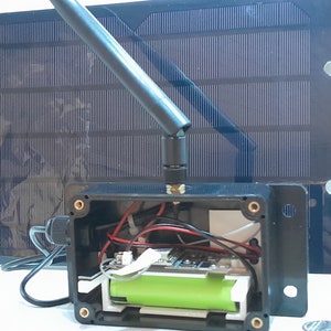 May include: A black plastic box with a green battery, wires, and a circuit board inside. A black antenna is attached to the top of the box. A black solar panel is attached to the side of the box.