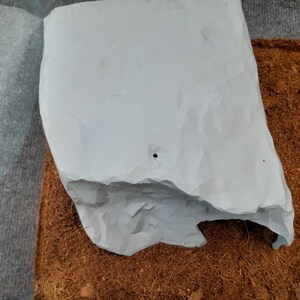 May include: A white ceramic planter with a rough, textured surface and a small hole in the center. The planter is sitting on a bed of brown coconut coir.