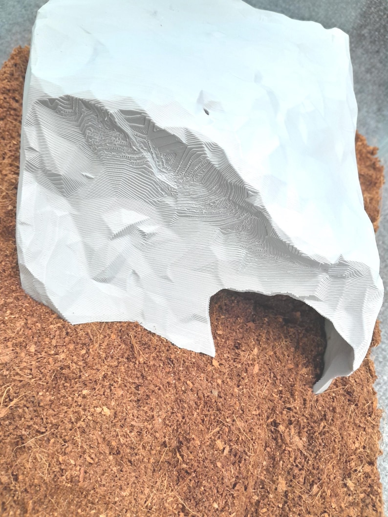 May include: A white 3D printed cave hide for small animals, sitting on a bed of brown coconut fiber.