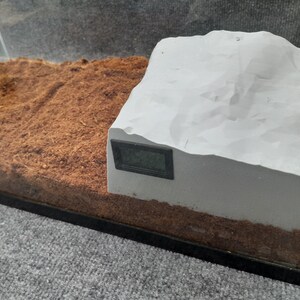 May include: A white, textured, rock-like structure with a black digital thermometer attached to the side. The structure is sitting on a bed of brown coconut fiber.