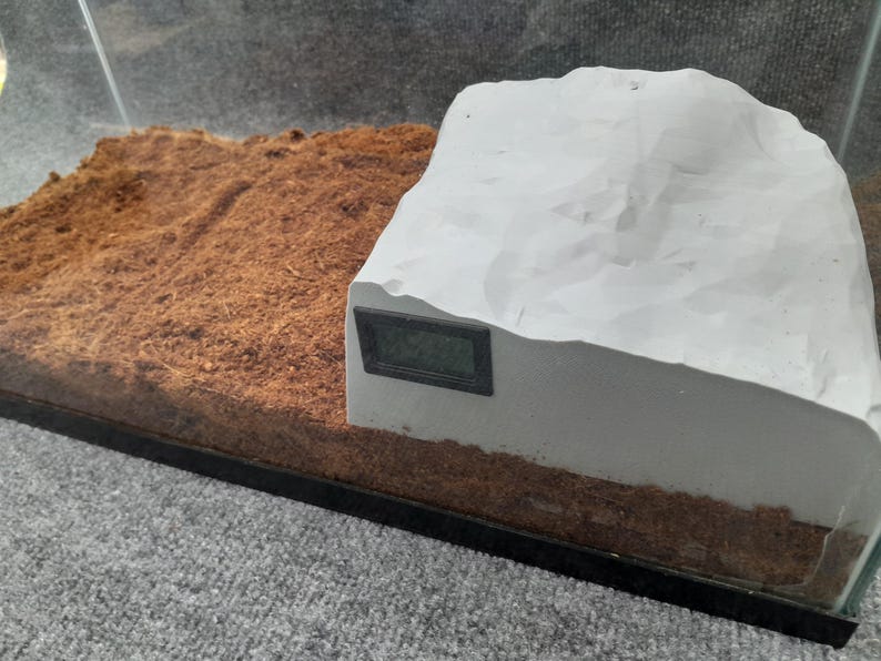 May include: A white, textured, rock-like structure with a black digital thermometer attached to the side. The structure is sitting on a bed of brown coconut fiber.