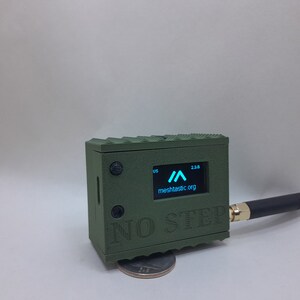 May include: A green, rectangular device with a small screen displaying a blue arrow and the text "meshtactic.org". The device has a black antenna and the text "NO STEP" printed on the front.