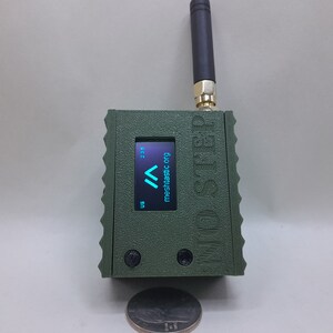 May include: A green, rectangular device with a small screen displaying the text "meshtactic.org" and a blue arrow pointing upwards. The device has a black antenna and the text "MIGHTY STEP" printed on the side.