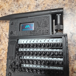 May include: A black, handheld electronic device with a small screen displaying "meshtastic.org". The device has a keyboard with black keys and white lettering. An antenna and a connector are attached to the top. The text "V2 HELTEC" is visible.